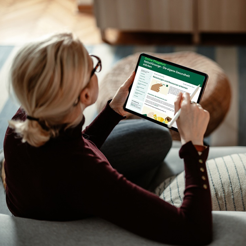 Frau sitzt auf dem Sofa und betrachtet auf einem Tablet einen Online-Pflegekurs